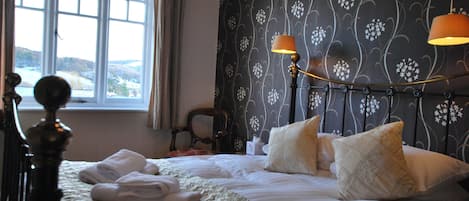 Una cama bien hecha con toallas dobladas, cabecero con lámparas fijadas a la pared y ventana con cortinas.