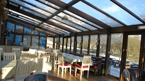 Un espace repas abrité, doté de tables et de chaises en bois, de grandes fenêtres et offrant une vue sur l’extérieur.