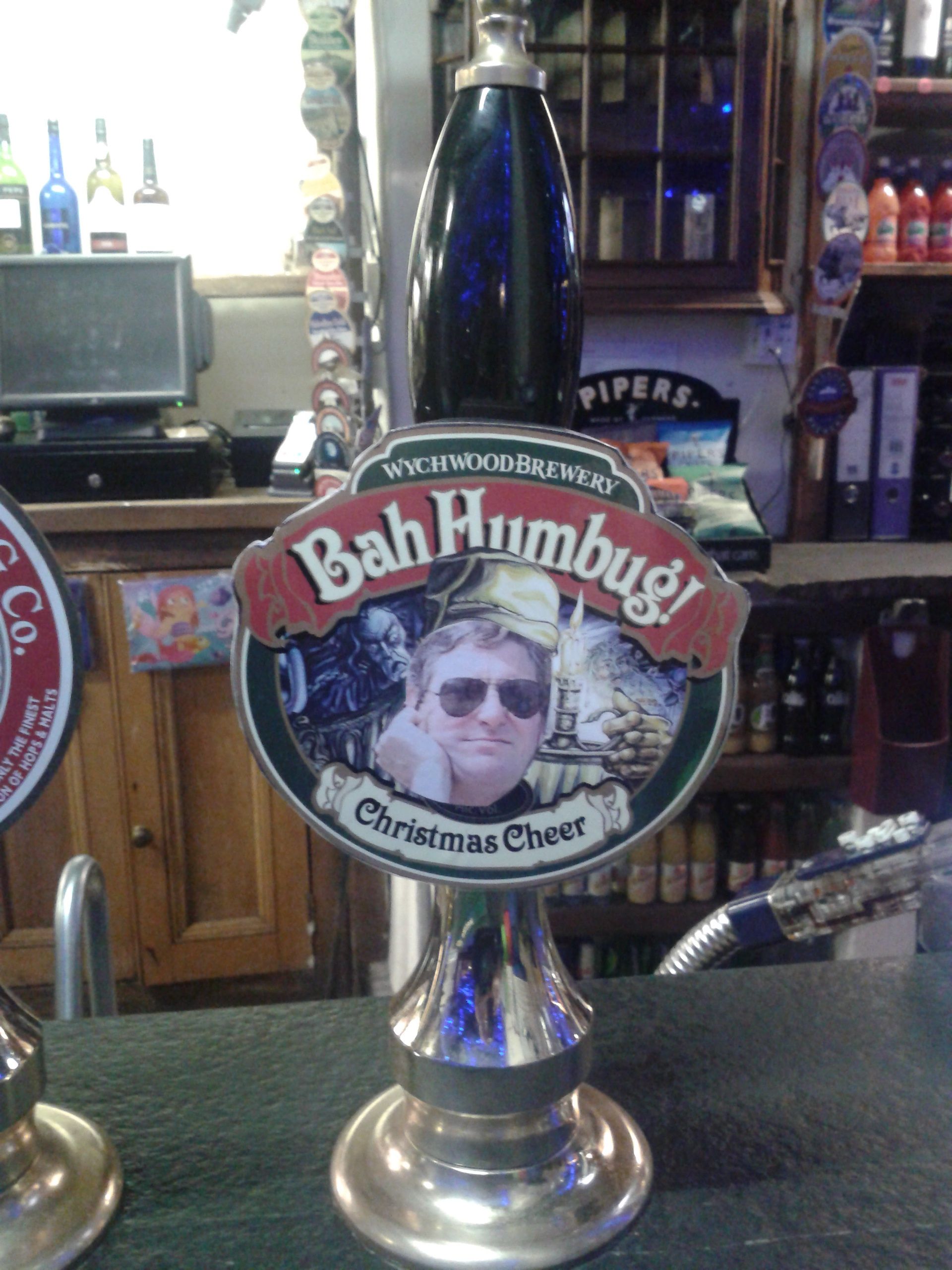 A beer tap labeled "Bah Humbug! Christmas Cheer" with a blurred face, surrounded by shelves of bottles and a computer monitor in the background.