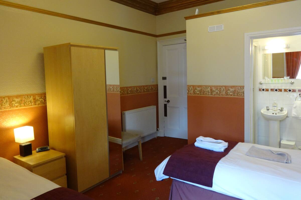 A hotel room with a bed, a nightstand, a wardrobe, a bathroom with a sink and mirror, and a door leading to another room.