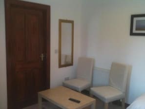A room with a wooden door, a mirror, two chairs, and a small table with a remote control.