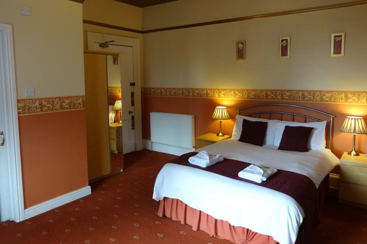 A hotel room with a bed, two bedside tables with lamps, a radiator, and a door leading to another room.