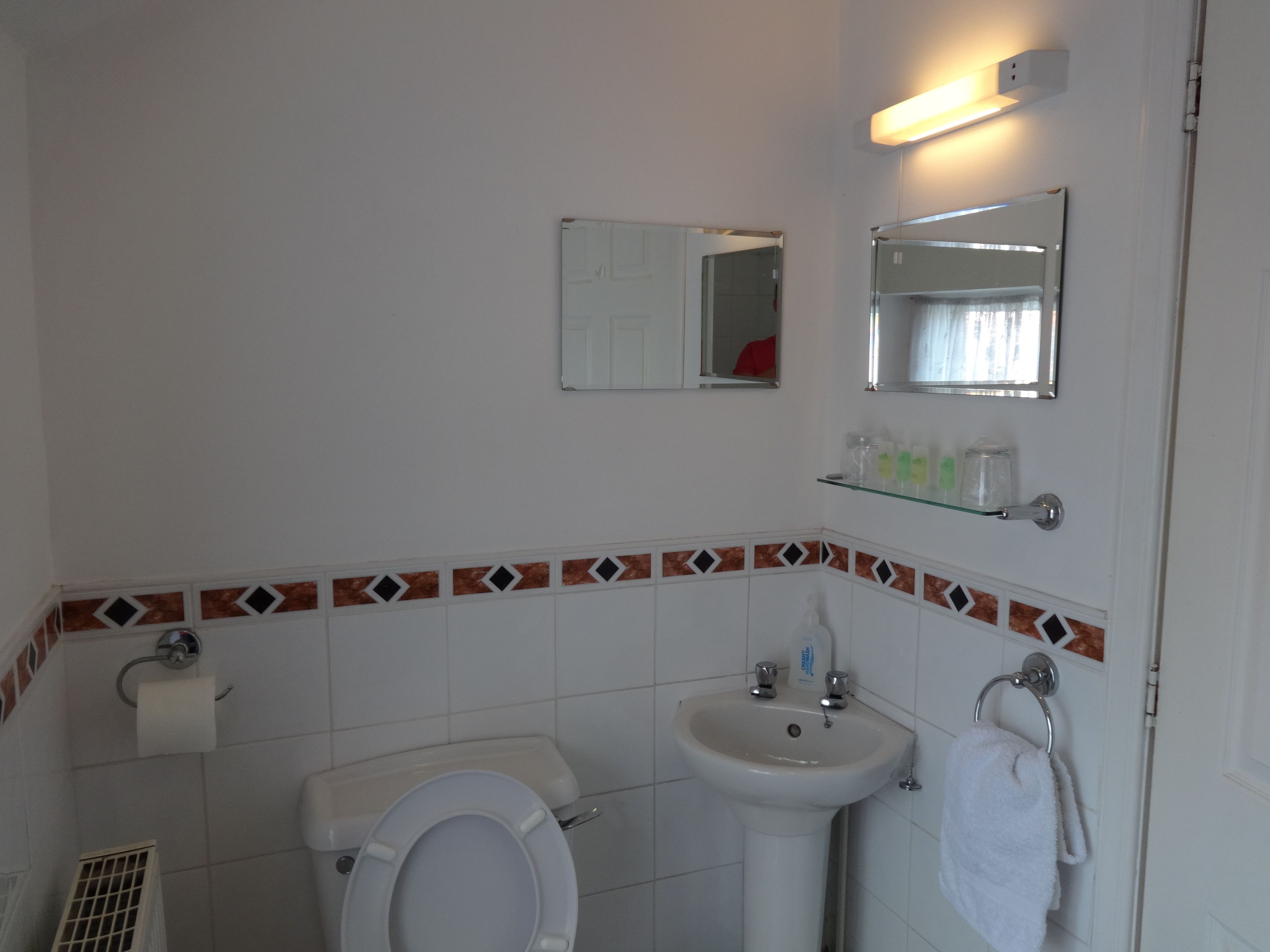 A bathroom with a toilet, sink, and mirrors. There is a towel rack and a shelf with toiletries.