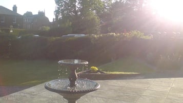 A fountain in a courtyard with a tiled floor and a grassy area with trees in the background.