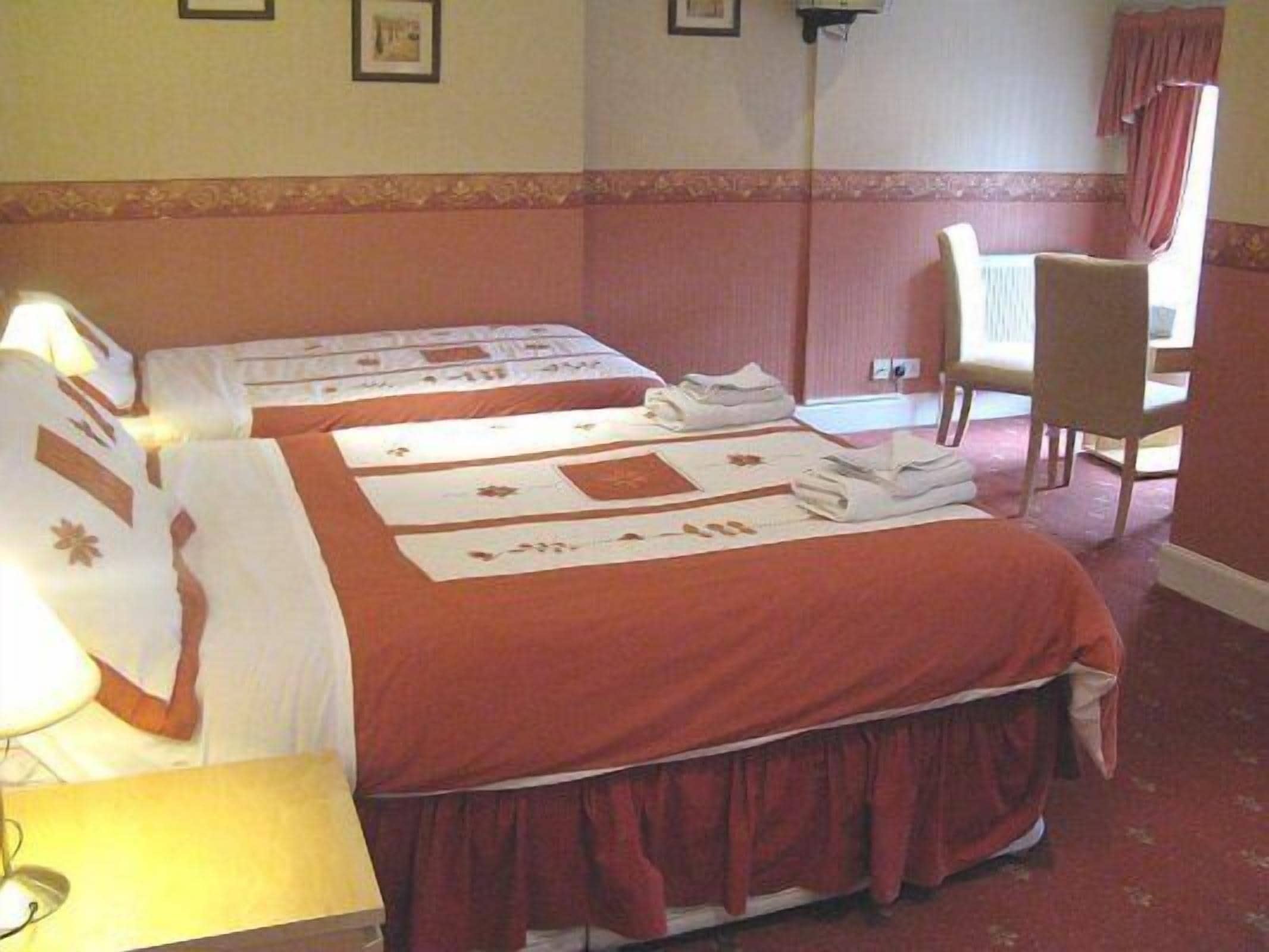 A hotel room with two beds, each with a red bedspread and white linens. There is a chair and a lamp in the room.