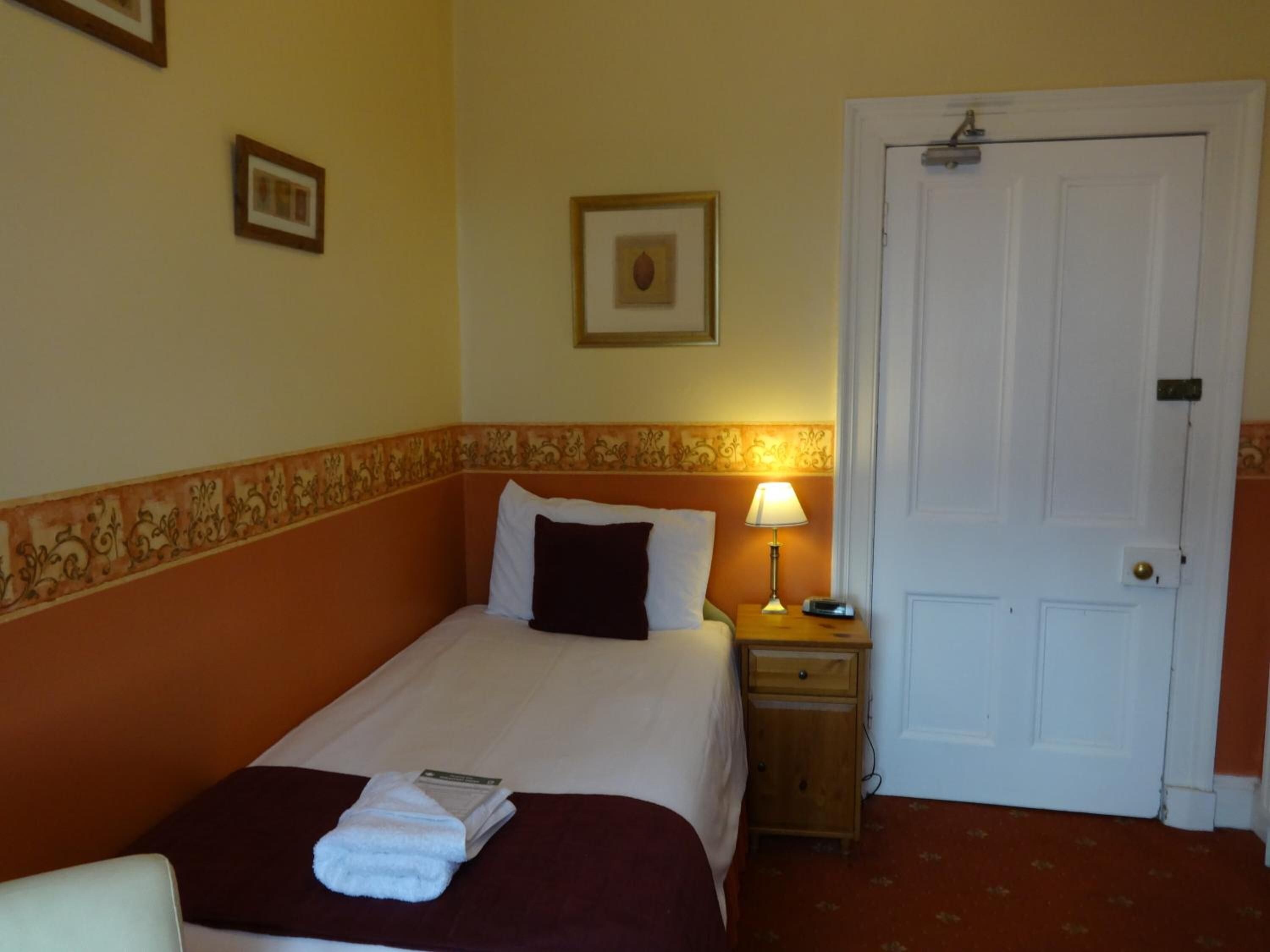 A hotel room with a bed, a nightstand with a lamp, a door, and a wall with a framed picture.