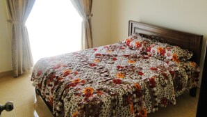 A bedroom with a bed covered in a floral bedspread, a wooden headboard, and a window with curtains. - BRAMA Servizez - Capital Park (Colombo)