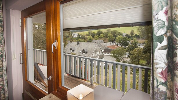 Un balcon doté d’une porte en bois, d’un banc et offrant une vue sur un quartier résidentiel avec des maisons et des arbres.