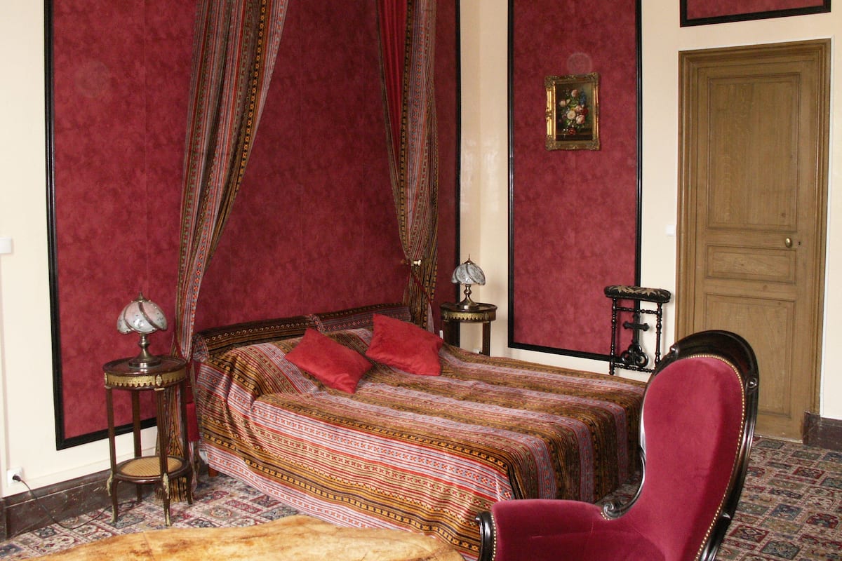 A bedroom with a four-poster bed, a red armchair, a small table with a lamp, and a wooden door.