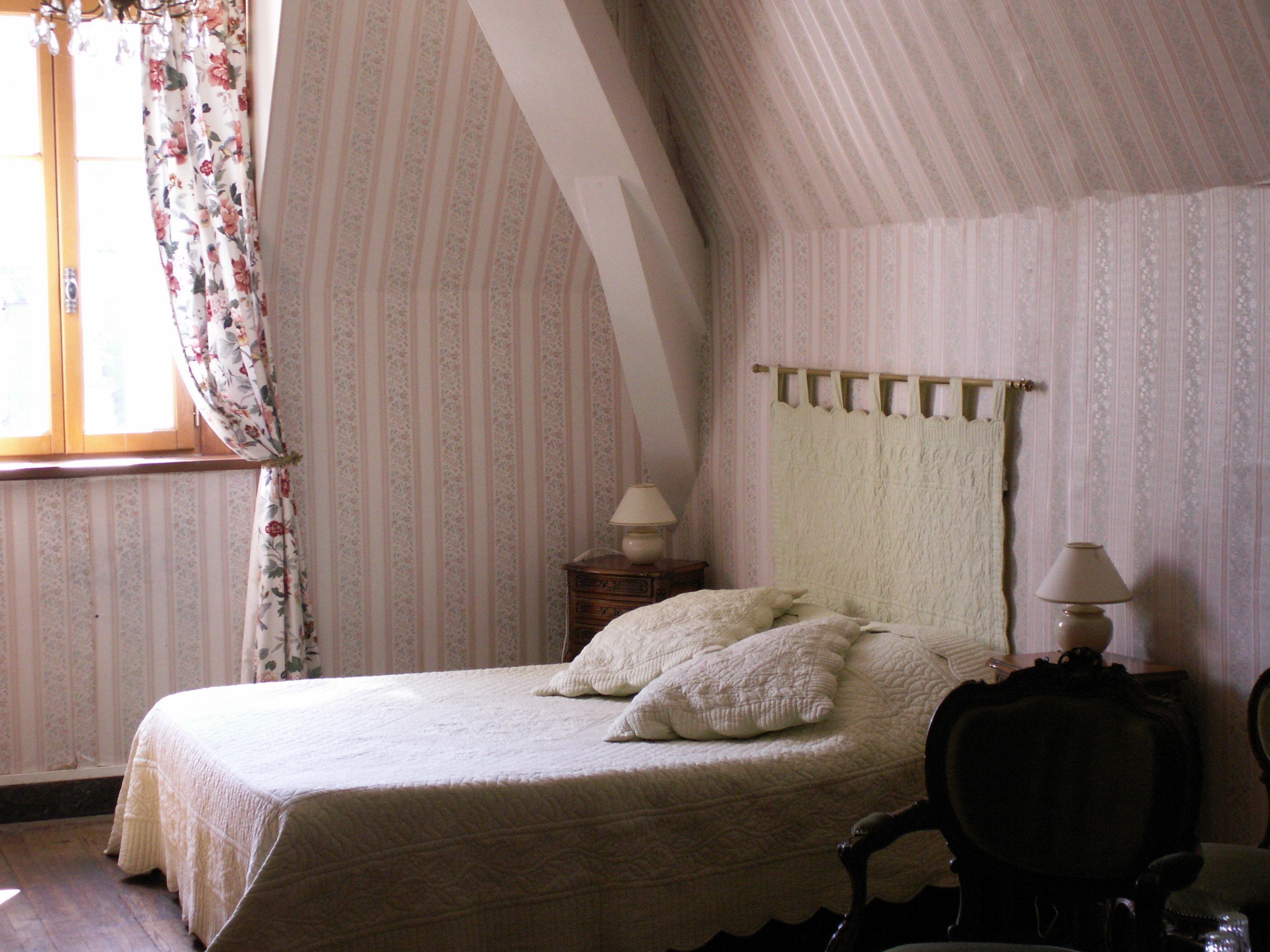 A bedroom with a large bed, two bedside lamps, a wooden chair, and a window with floral curtains.