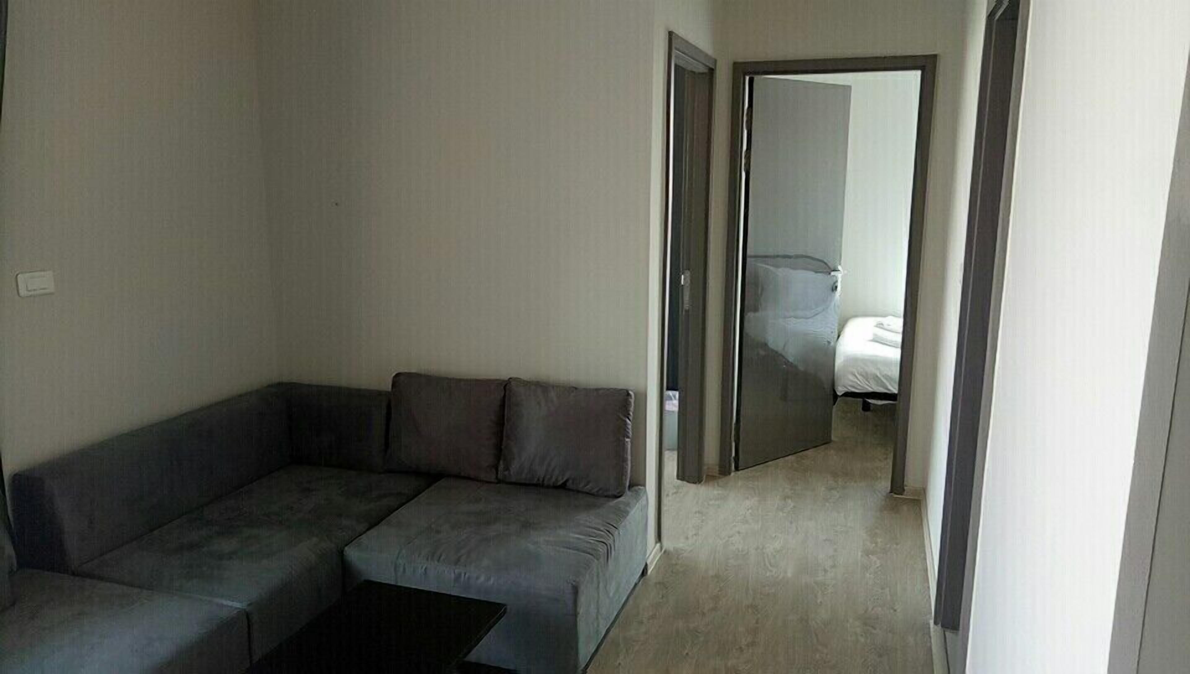 A modern, minimalist living room with a grey sectional sofa, a small coffee table, and a visible bedroom with a bed and a nightstand.