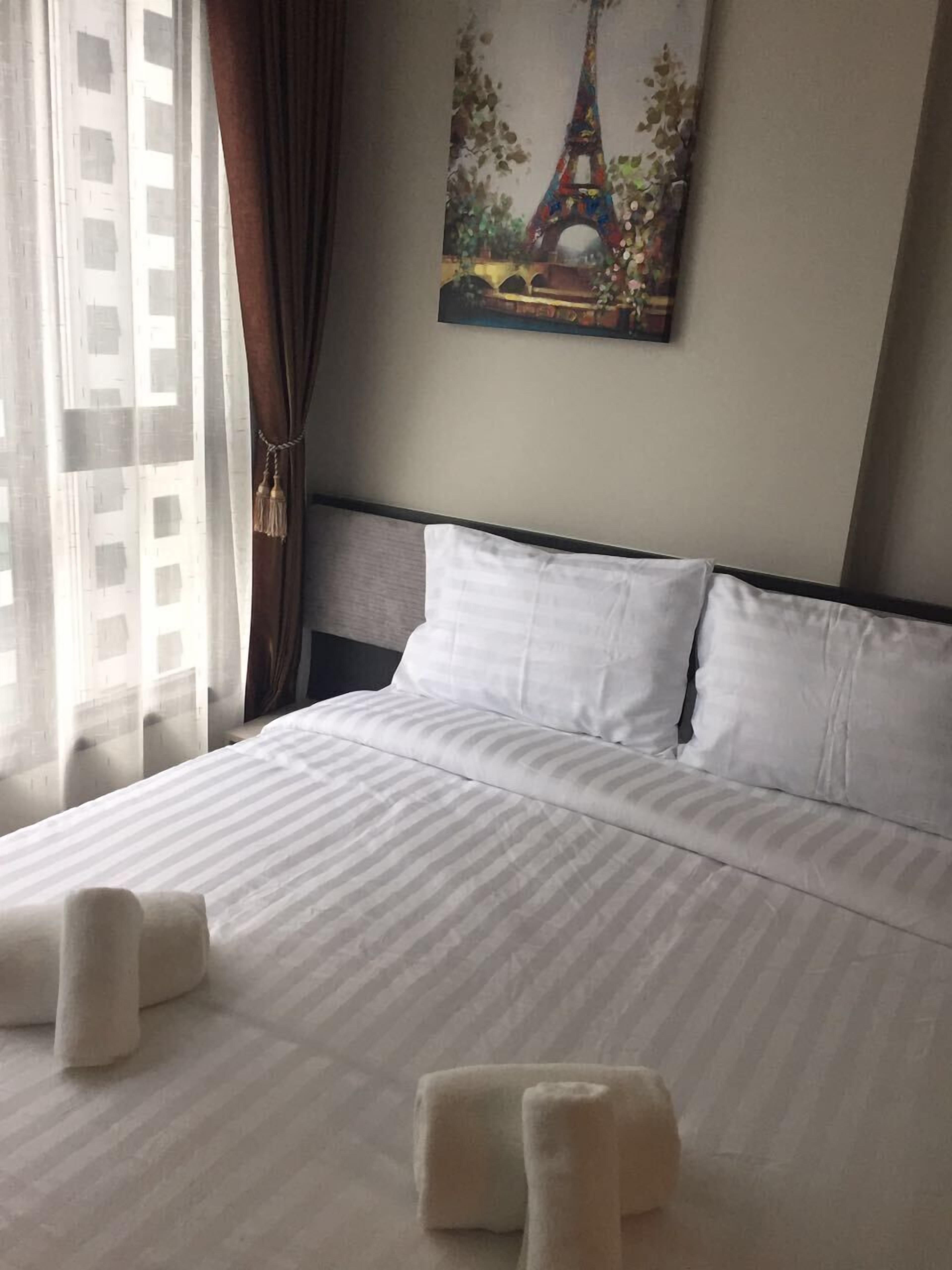 Un lit bien fait, avec du linge de lit blanc et des oreillers, un tableau représentant la Tour Eiffel accroché au mur, et une fenêtre avec des rideaux.