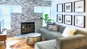 A living room with a stone wall, a fireplace, a grey sofa, a round ottoman, and framed pictures on the wall. - Walnut Mews - Quiet Creekside townhome next to Pearl Street (Boulder)