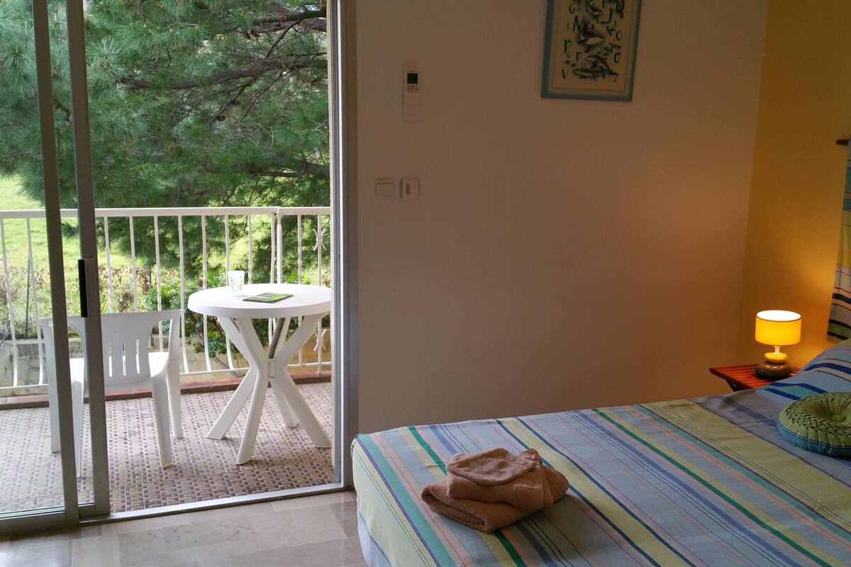 A bedroom with a bed, a bedside table with a lamp, a window with a view of greenery, and a balcony with a table and chairs.
