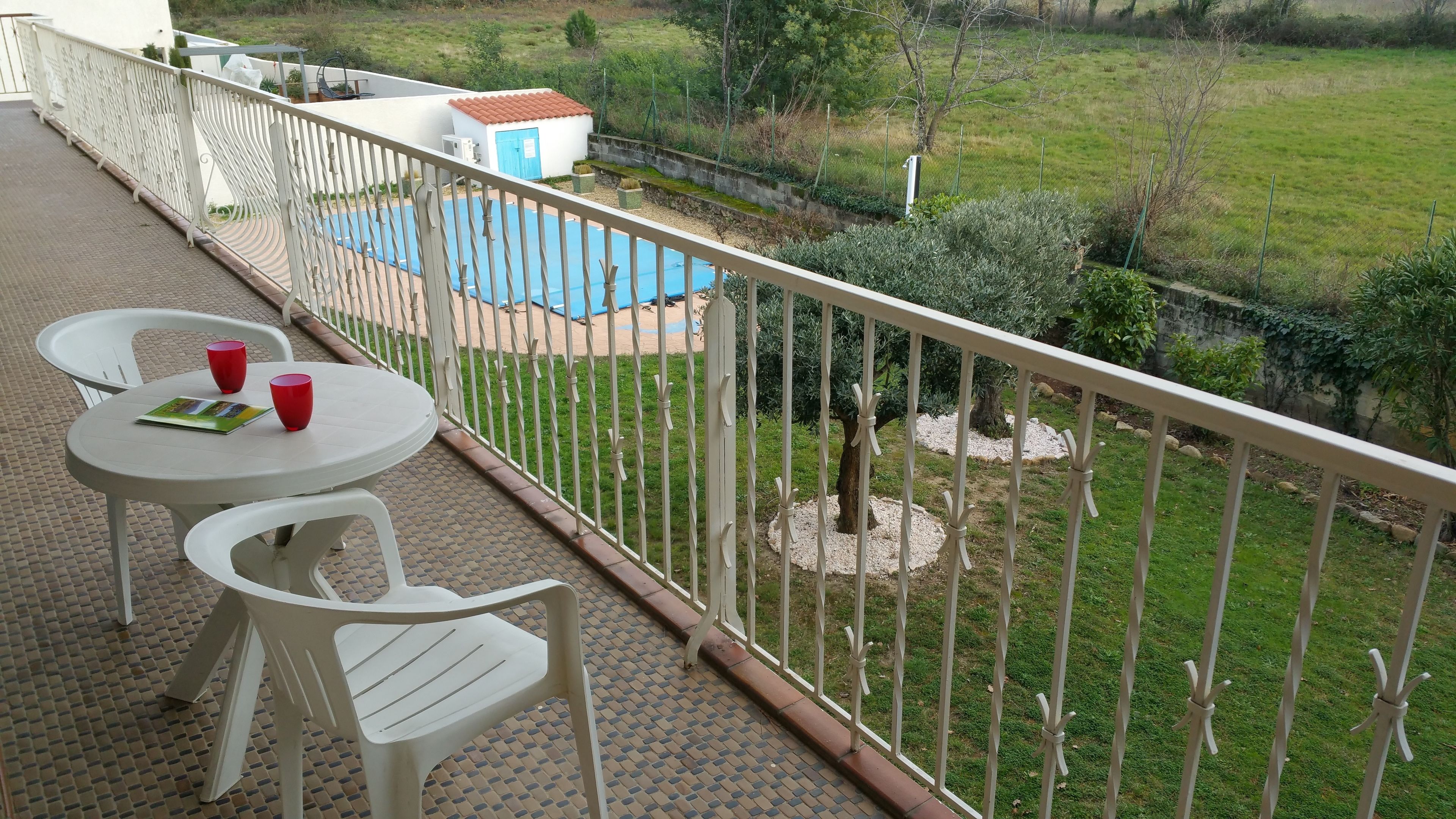 A balcony with two white plastic chairs and a table, overlooking a swimming pool and a garden.