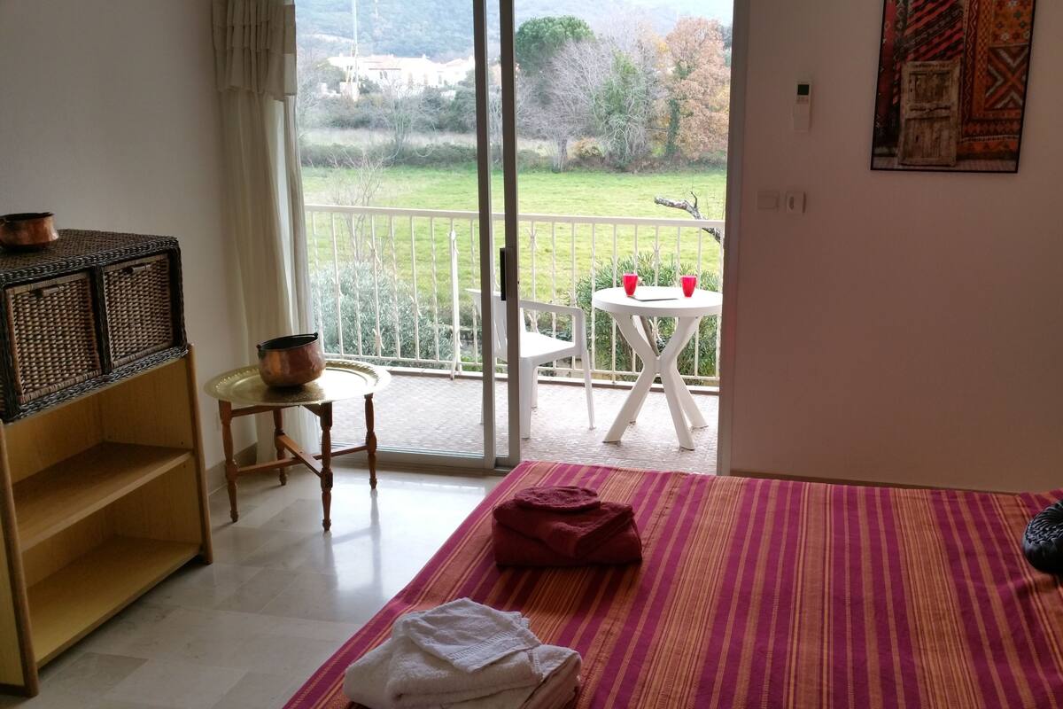 A hotel room with a bed, a small table with chairs, a balcony with a table and chairs, and a view of the outdoors.