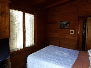 Ein Zimmer in einer Holzhütte mit einem Bett, einem Fernseher und einem Fenster mit Vorhängen.
