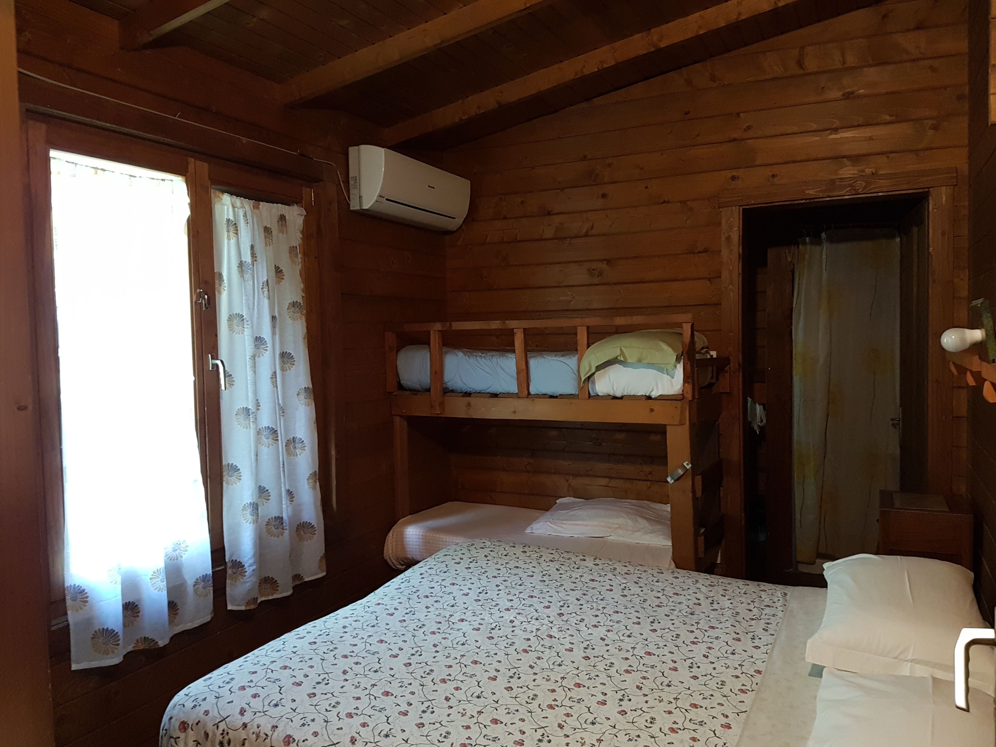 A wooden cabin room with bunk beds, a window with curtains, and an air conditioning unit.
