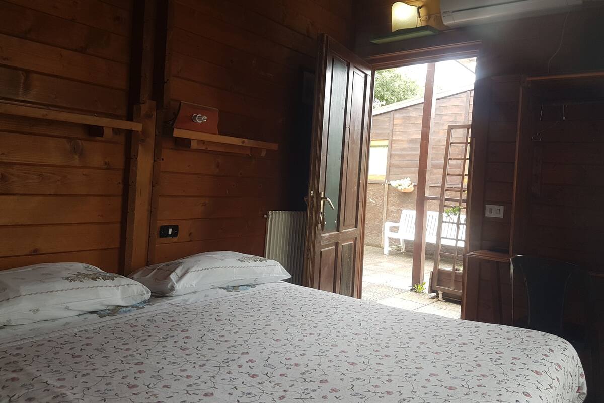 Ein Zimmer mit Holzboden, einem Bett, einem Heizkörper, einem Regal und einer offenen Tür, die nach draußen führt.