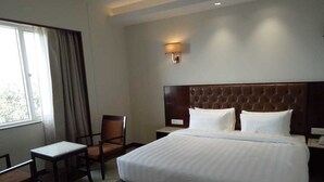 A hotel room with a large bed, a bedside table, a chair, and a window with curtains. - Hotel Parador (Firozabad)