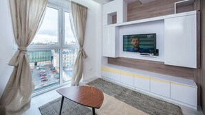 A modern hotel room with a large window, a flat-screen TV mounted on the wall, a small wooden table, and a patterned rug.