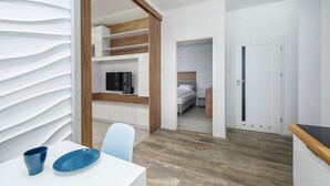 A modern hotel room with a wooden floor, white walls, a TV on a wooden shelf, a bed with a headboard, and a door leading to another room.