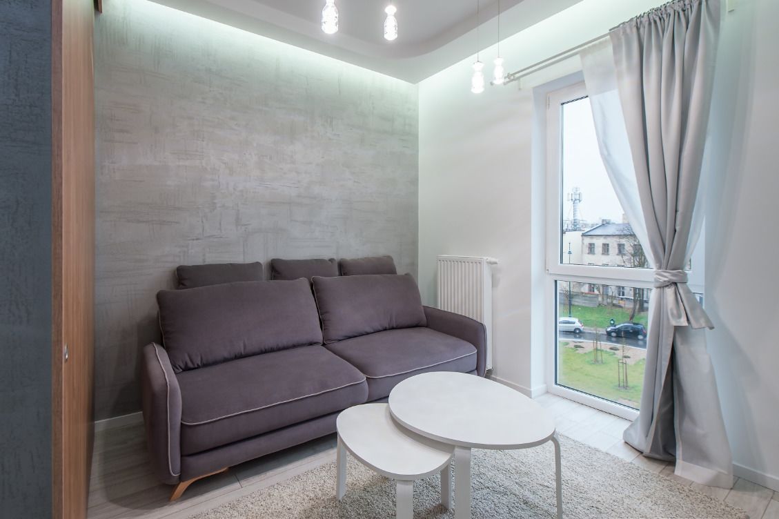 Un soggiorno moderno con un divano grigio, due tavolini bianchi, un'ampia finestra con tende e vista su un edificio e una strada.