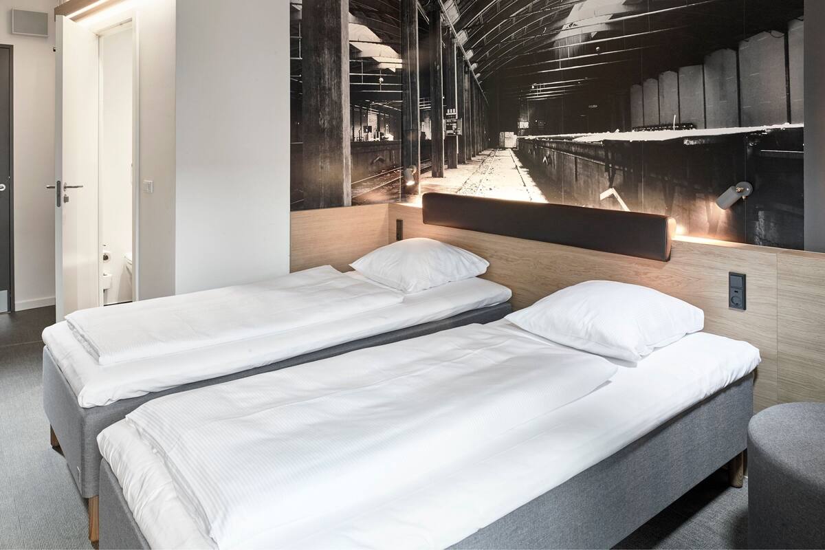 Une chambre d’hôtel avec deux lits simples, une tête de lit en bois, une grande fenêtre donnant sur une gare et un petit coin salon.