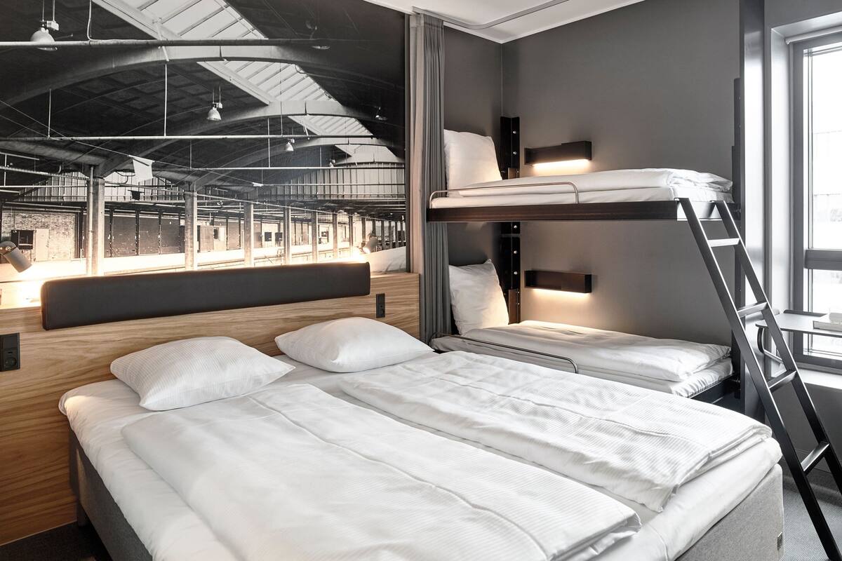 Une chambre d’hôtel moderne avec un lit superposé, une tête de lit en bois, un bureau avec une chaise et une grande fenêtre donnant sur l’espace intérieur.