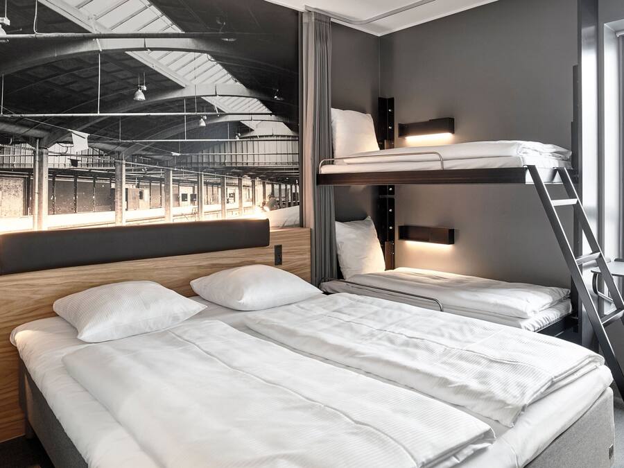 Une chambre d’hôtel moderne avec un lit superposé, une tête de lit en bois, un bureau avec une chaise et une grande fenêtre donnant sur l’espace intérieur.