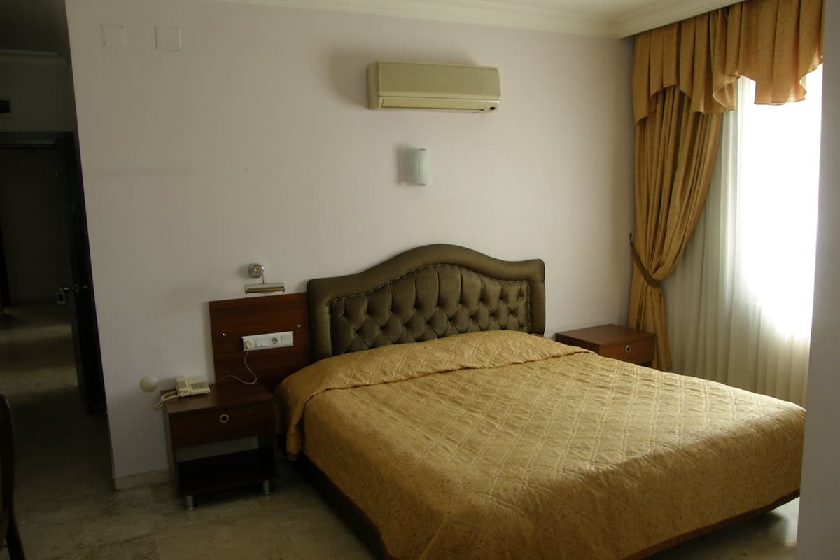 A hotel room with a large bed, a bedside table with a lamp, a wall-mounted air conditioner, and a window with curtains.