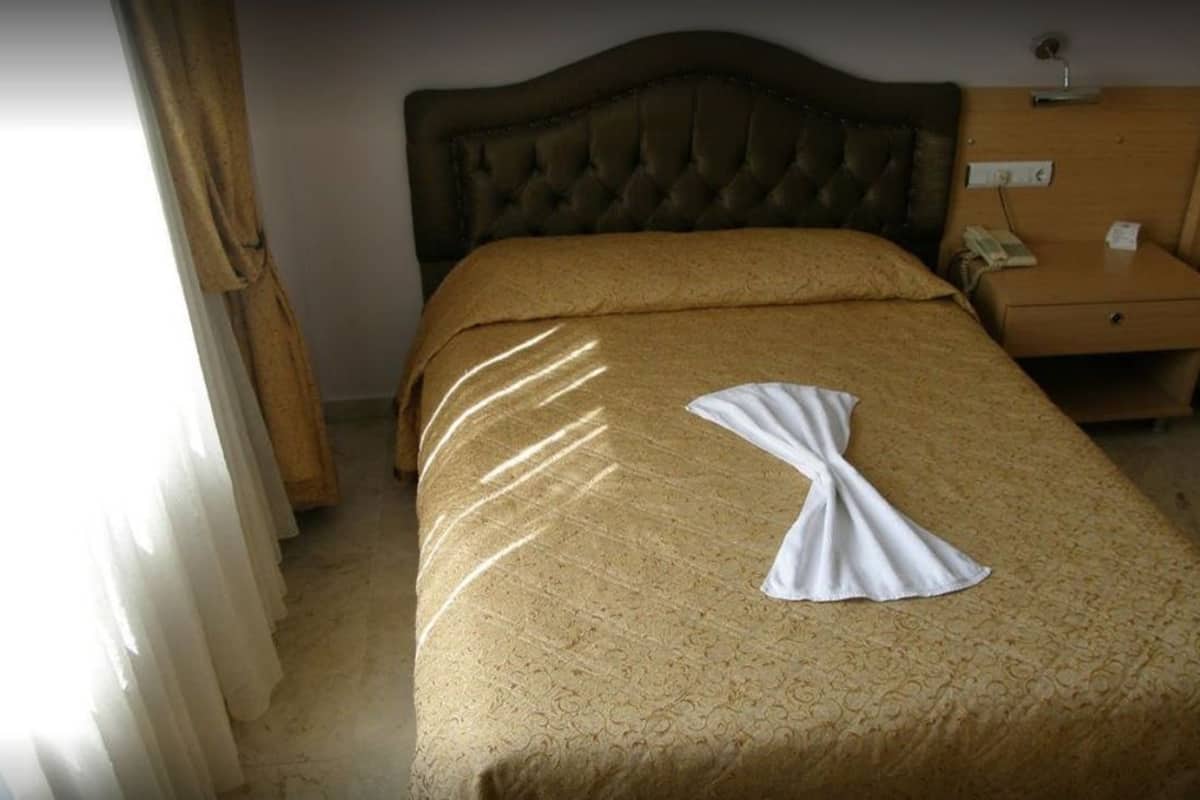 A hotel room with a large bed, a wooden headboard, a nightstand with a lamp, and a window with curtains.