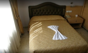 Habitación de hotel con una cama grande, cabecera de madera, mesita de noche con lámpara y una ventana con cortinas.