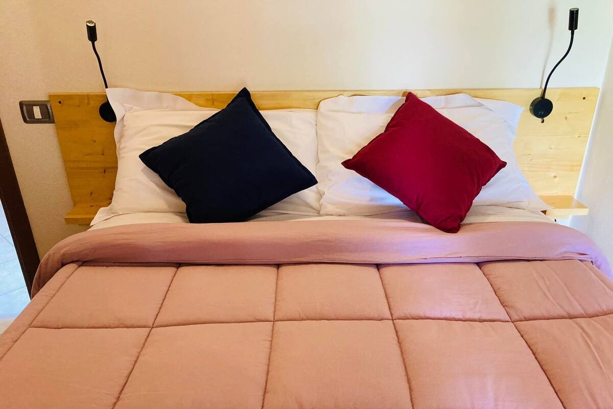 A bed with a pink quilted cover, two pillows (one black, one red), and a wooden headboard with a power outlet.