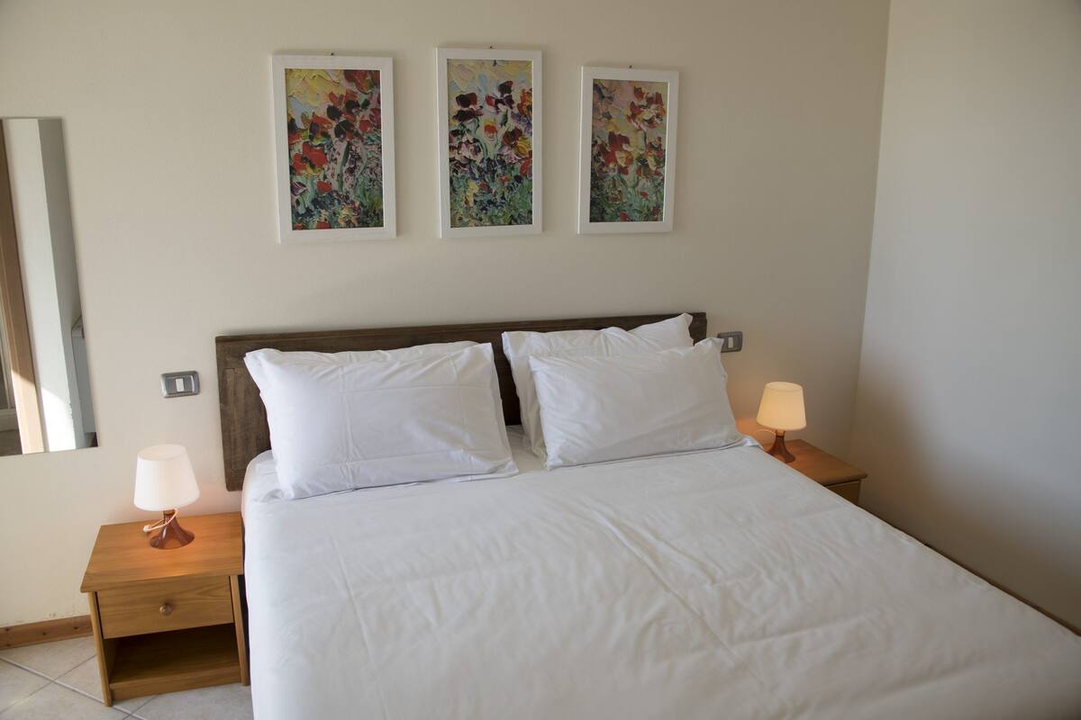 A neatly made bed with white linens, two bedside tables with lamps, and three floral paintings on the wall.