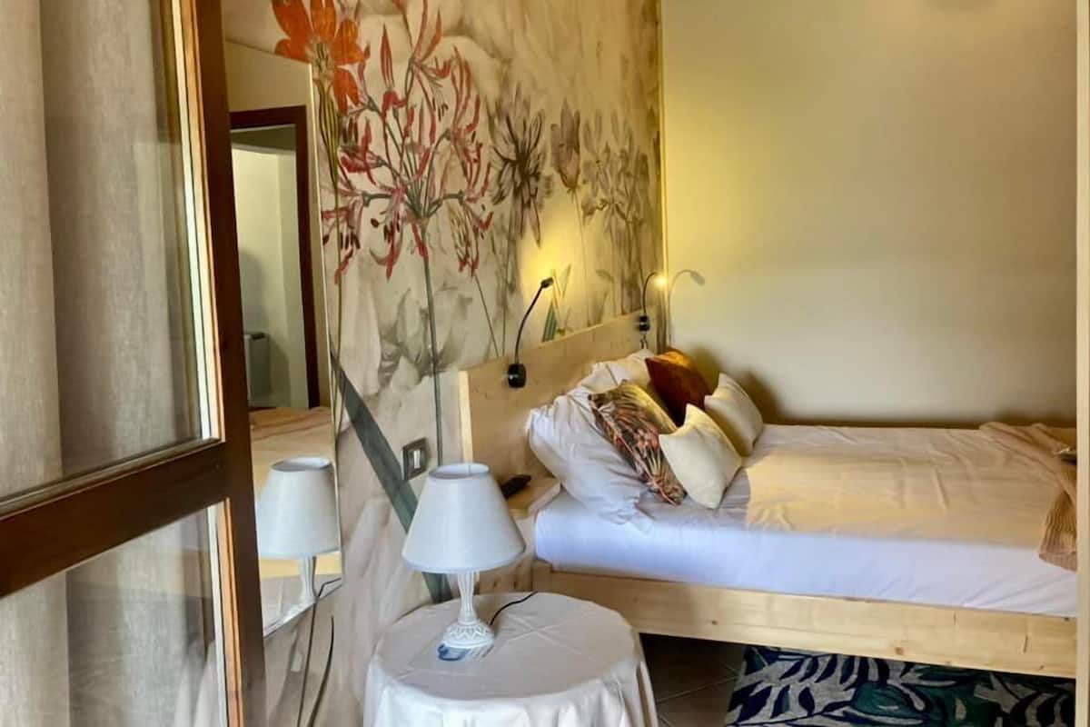 A hotel room with a bed, a bedside table with a lamp, a wall with a floral mural, and a mirror.