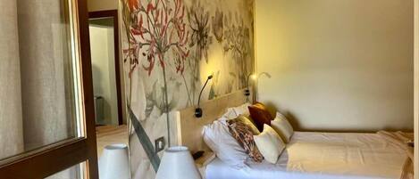 A hotel room with a bed, a bedside table with a lamp, a wall with a floral mural, and a mirror.