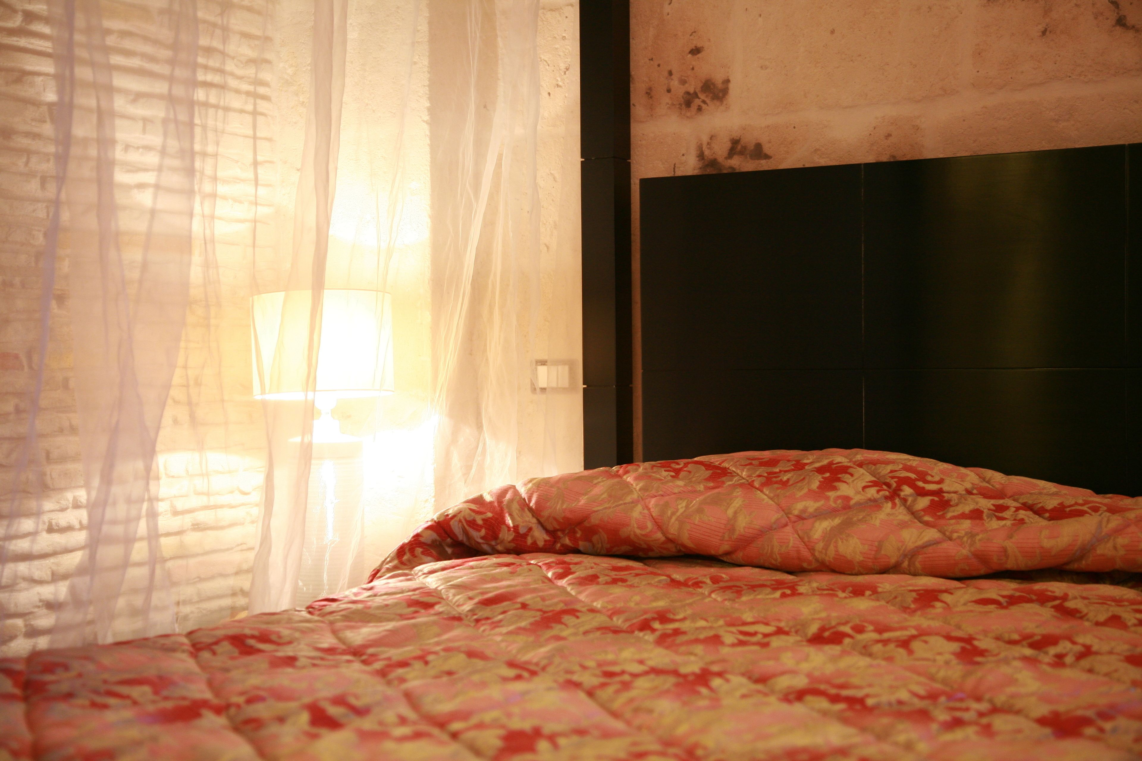 A bed with a patterned bedspread, a bedside lamp, and sheer curtains.
