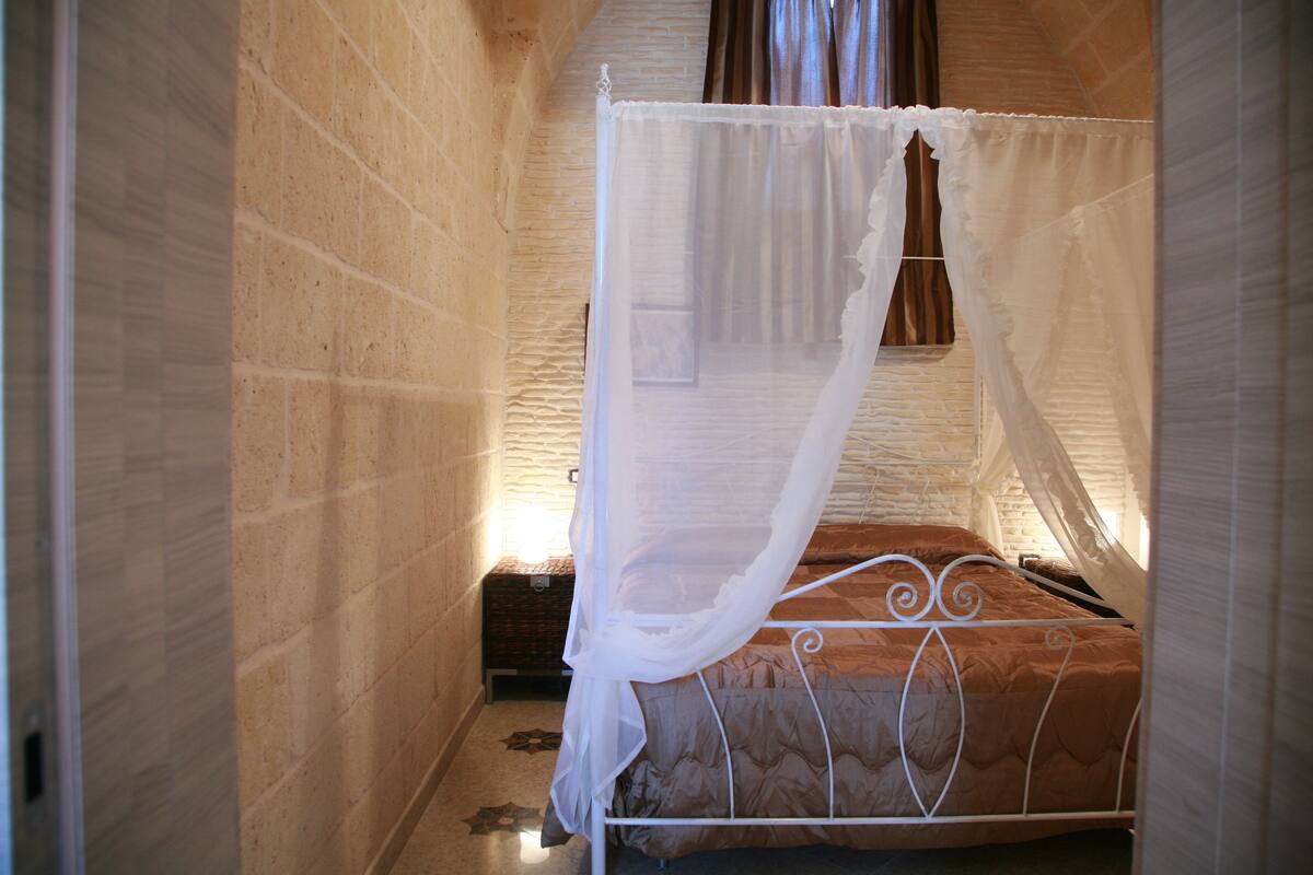 A single bed with a white metal frame and a brown quilt, a white canopy, and a small wooden chest beside it.