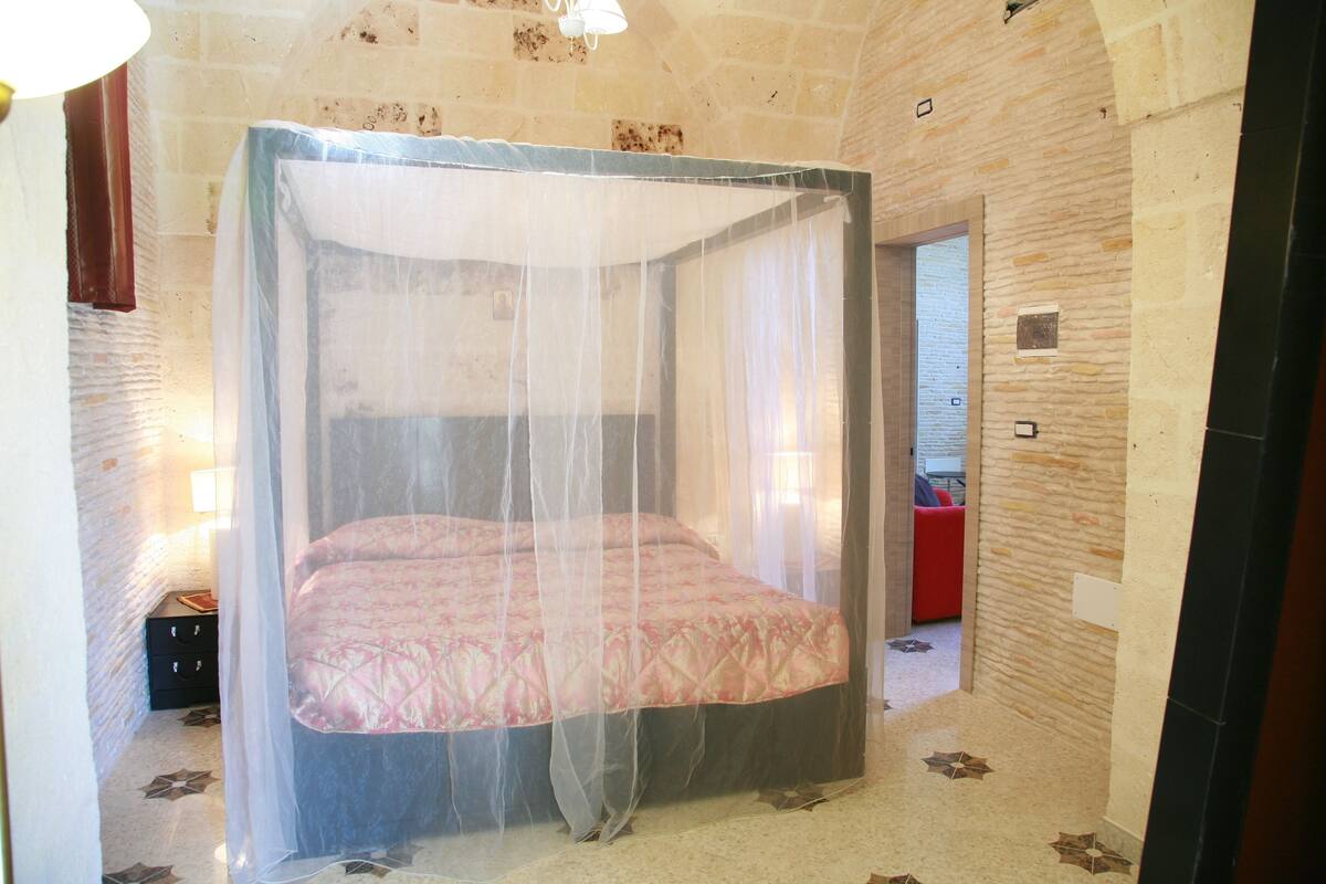 A four-poster bed with a sheer canopy, a red chair, and a stone wall.