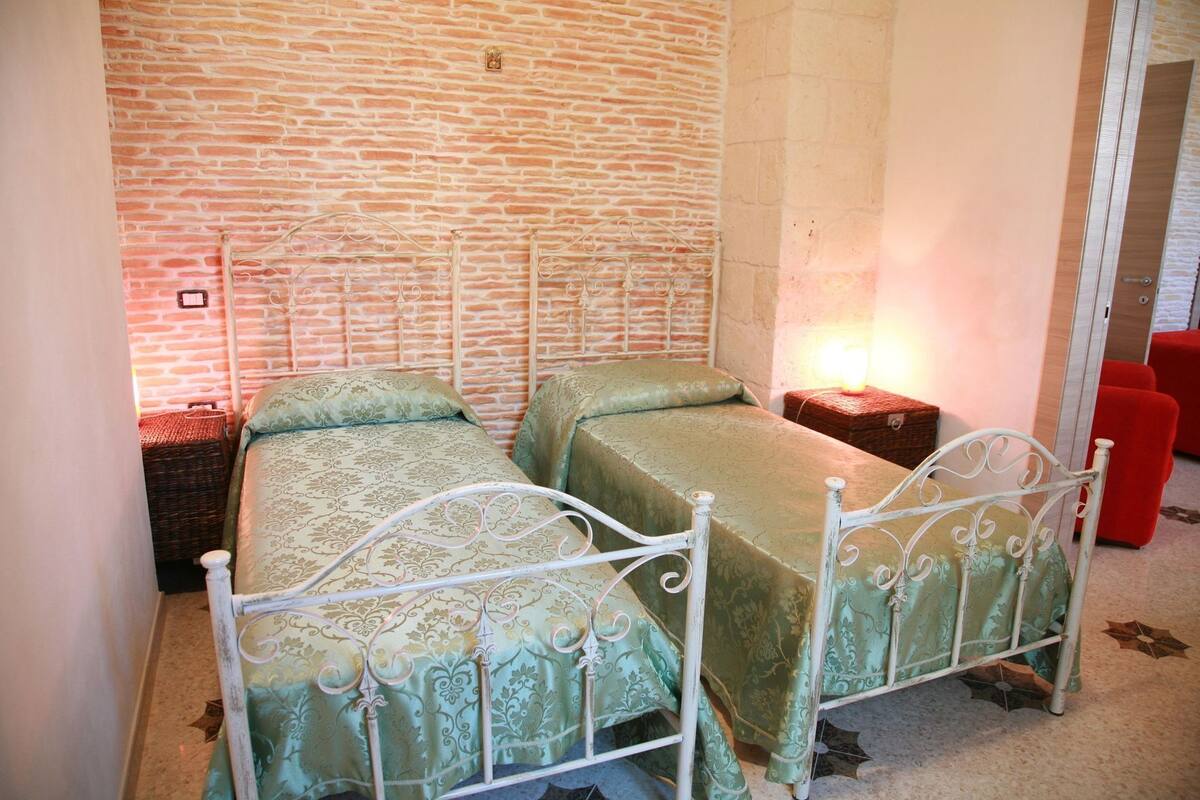 A room with two metal beds, a brick wall, a small table with a lamp, and a red chair.