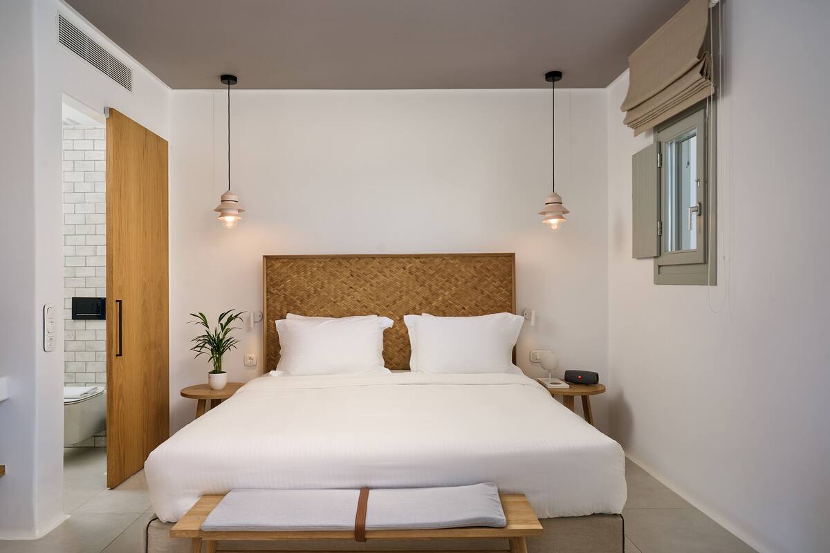 Un dormitorio moderno con cabecera de madera, cama grande, mesitas de noche, una planta en maceta y un ventanal con cortinas.