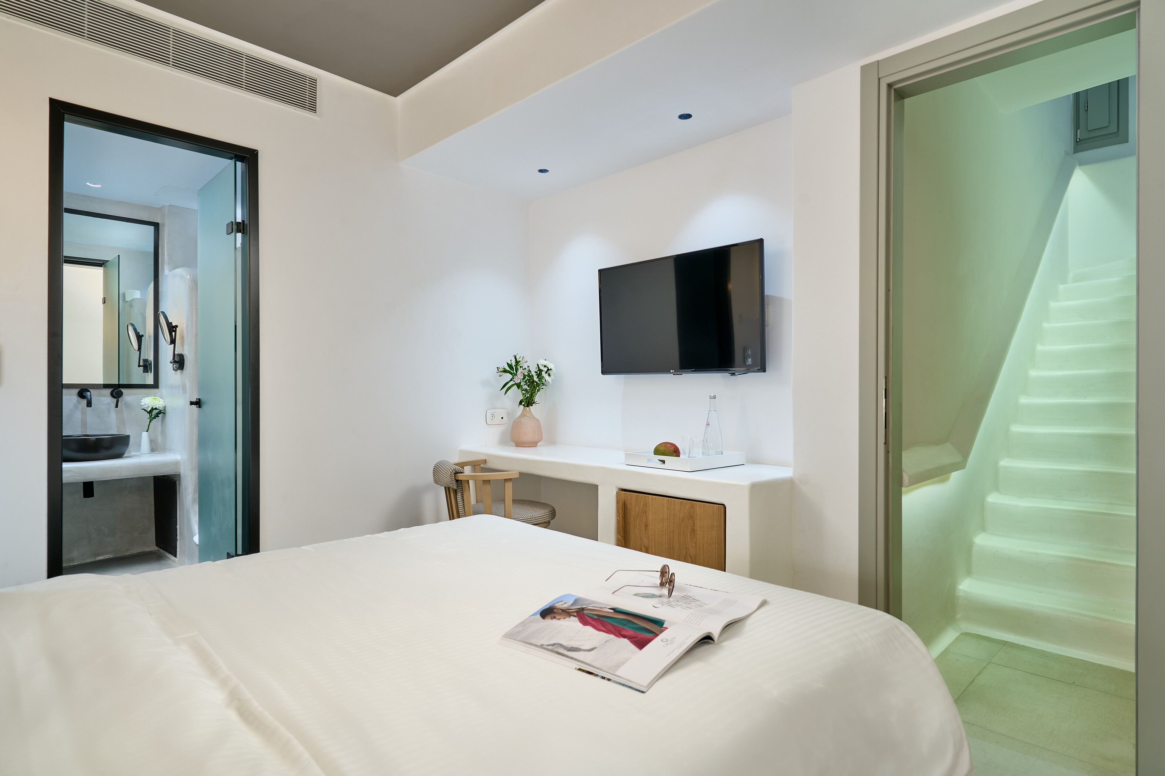 A modern hotel room with a bed, a desk with a flat-screen TV, a bathroom with a sink and mirror, and a staircase.