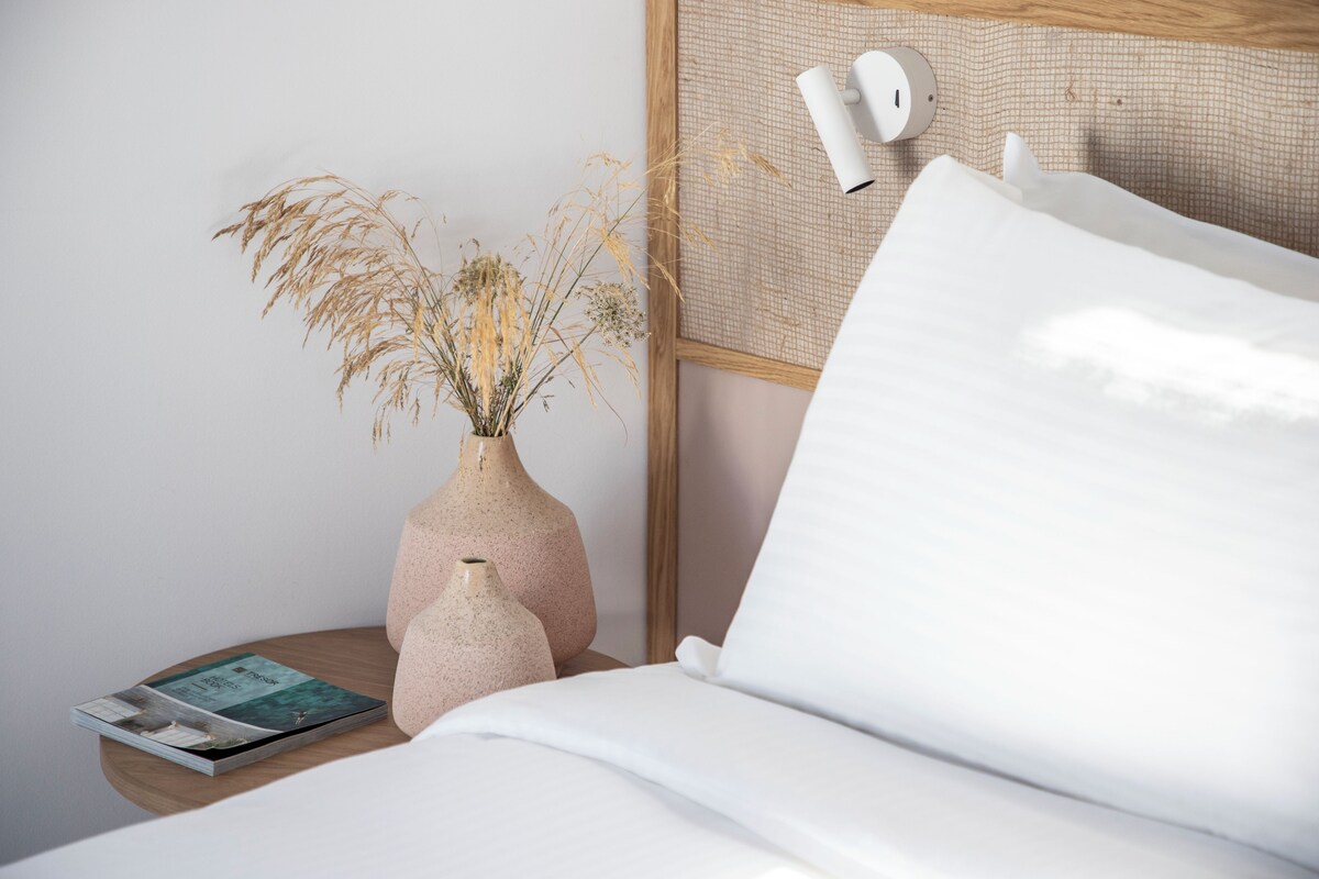 Un lit bien fait, avec du linge de lit blanc, une table de chevet en bois ornée d’un vase contenant des plantes séchées et un magazine.