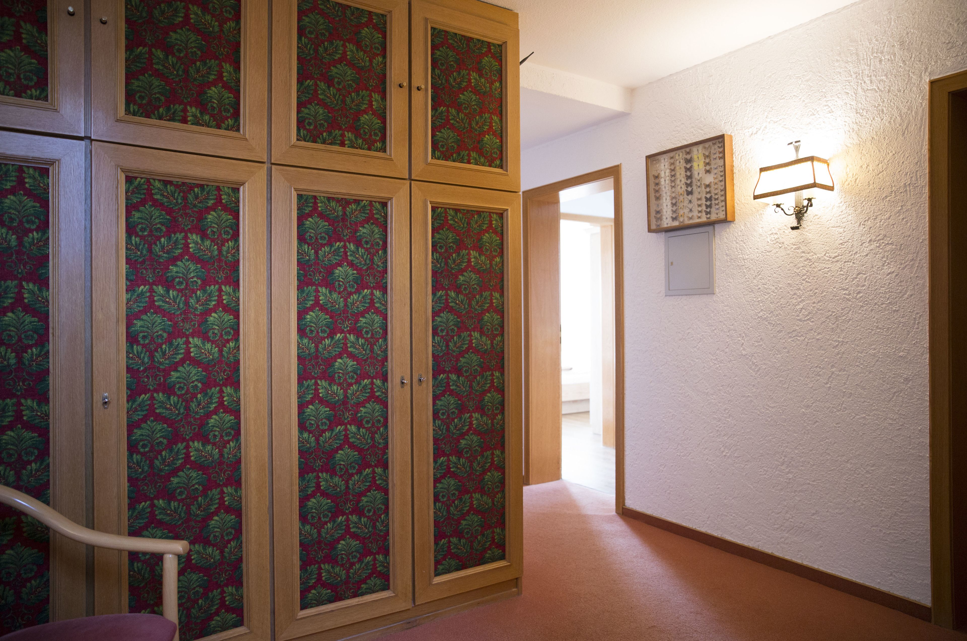 A hallway with a patterned wallpaper, a wooden cabinet, a wall-mounted light fixture, and a door leading to another room.