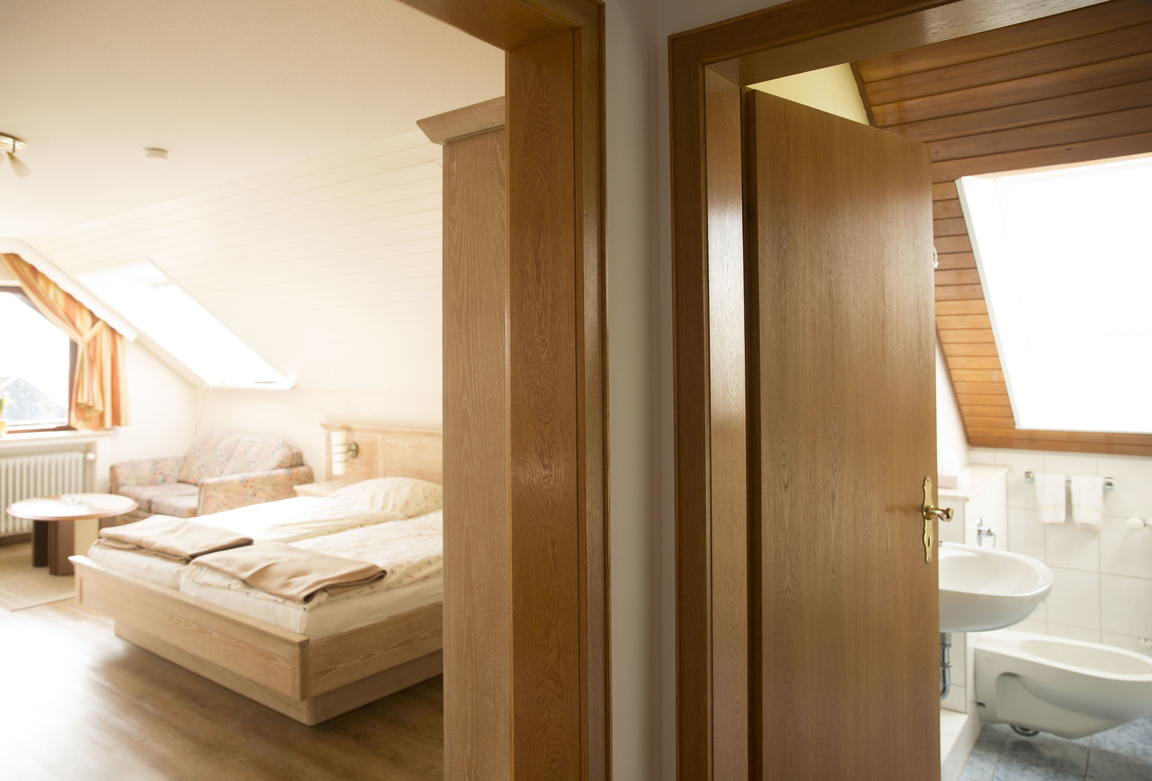 Ein Schlafzimmer mit Bett, Nachttisch, einem Fenster mit Vorhängen und Blick auf ein Badezimmer mit Waschbecken und Badewanne.