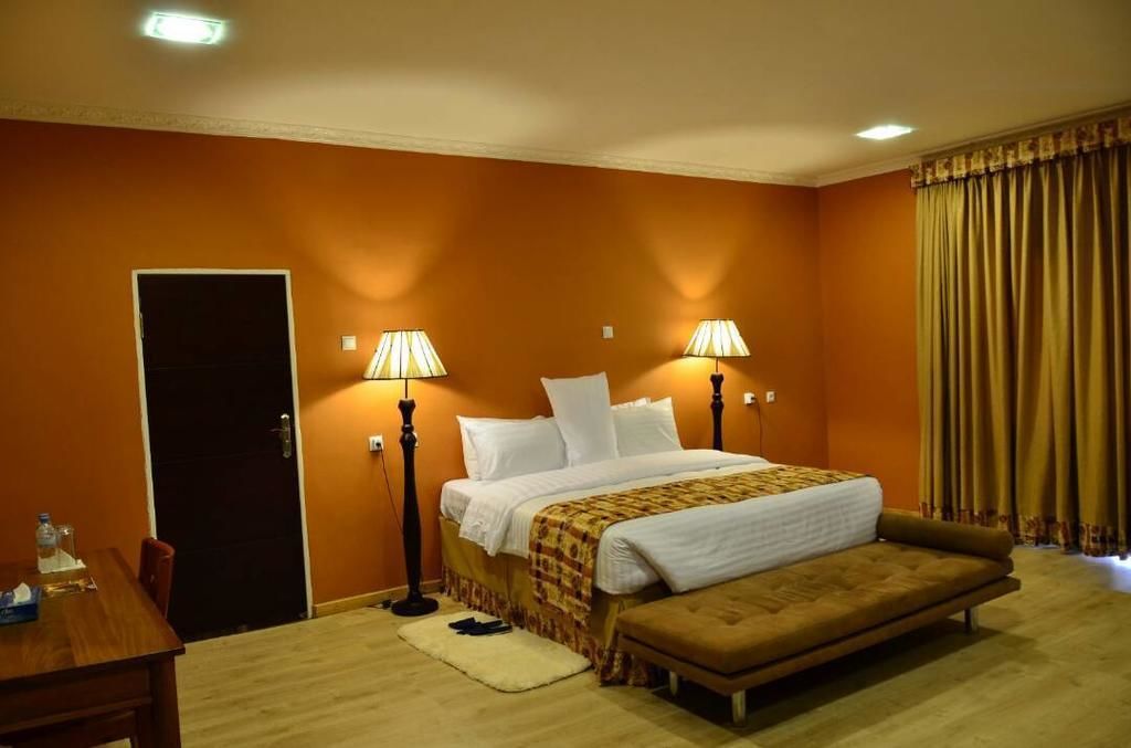 A hotel room with a bed, bedside lamps, a bench, a table with items, and a door.