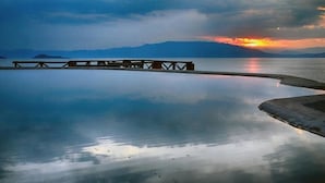 Ruhiges Gewässer mit einer hölzernen Mole, die sich ins Wasser erstreckt, umgeben von Bergen und einem bewölkten Himmel.
