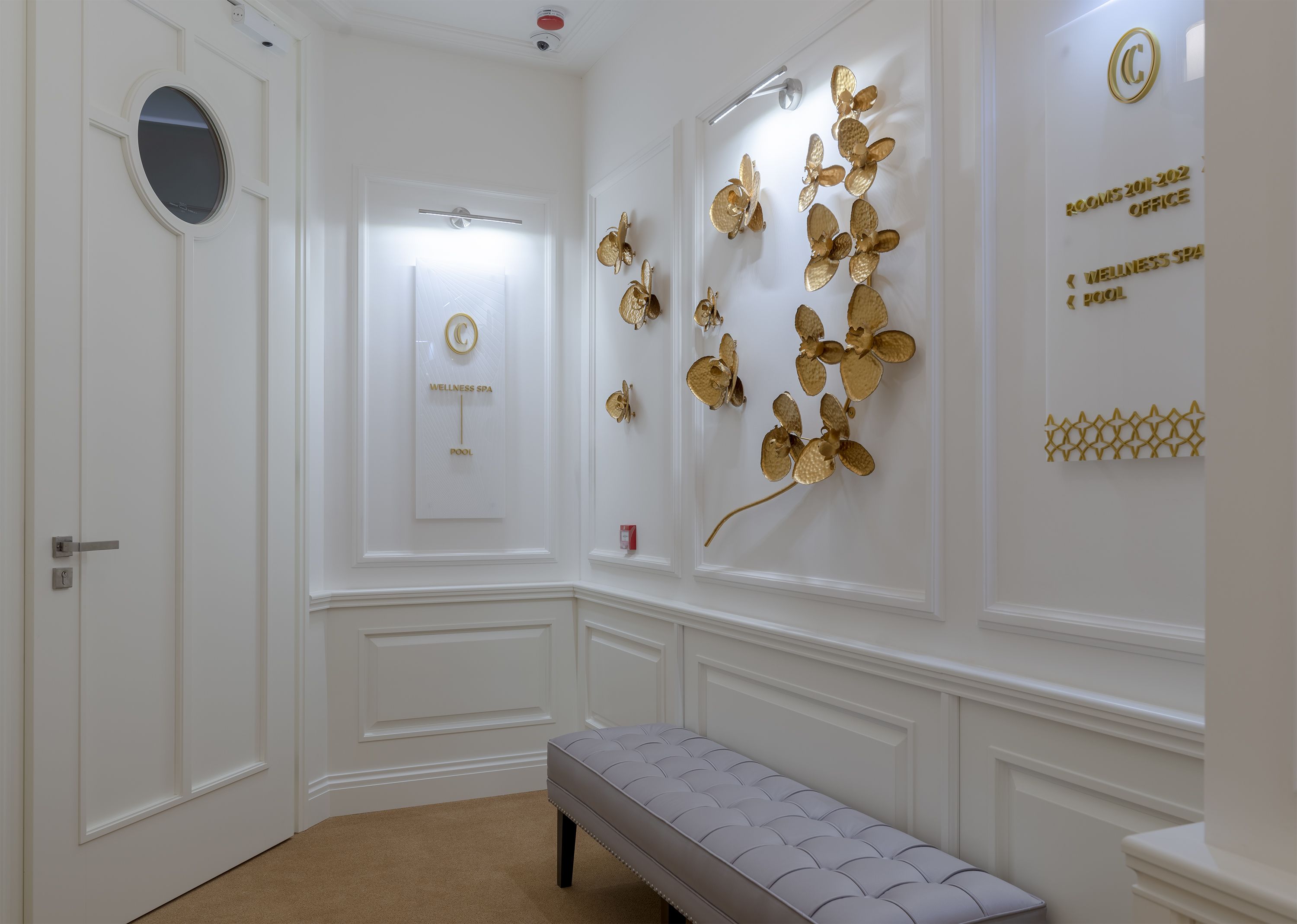 A hallway with a bench, a door with a circular mirror, a wall-mounted sign indicating a wellness spa and pool, and decorative gold elements.