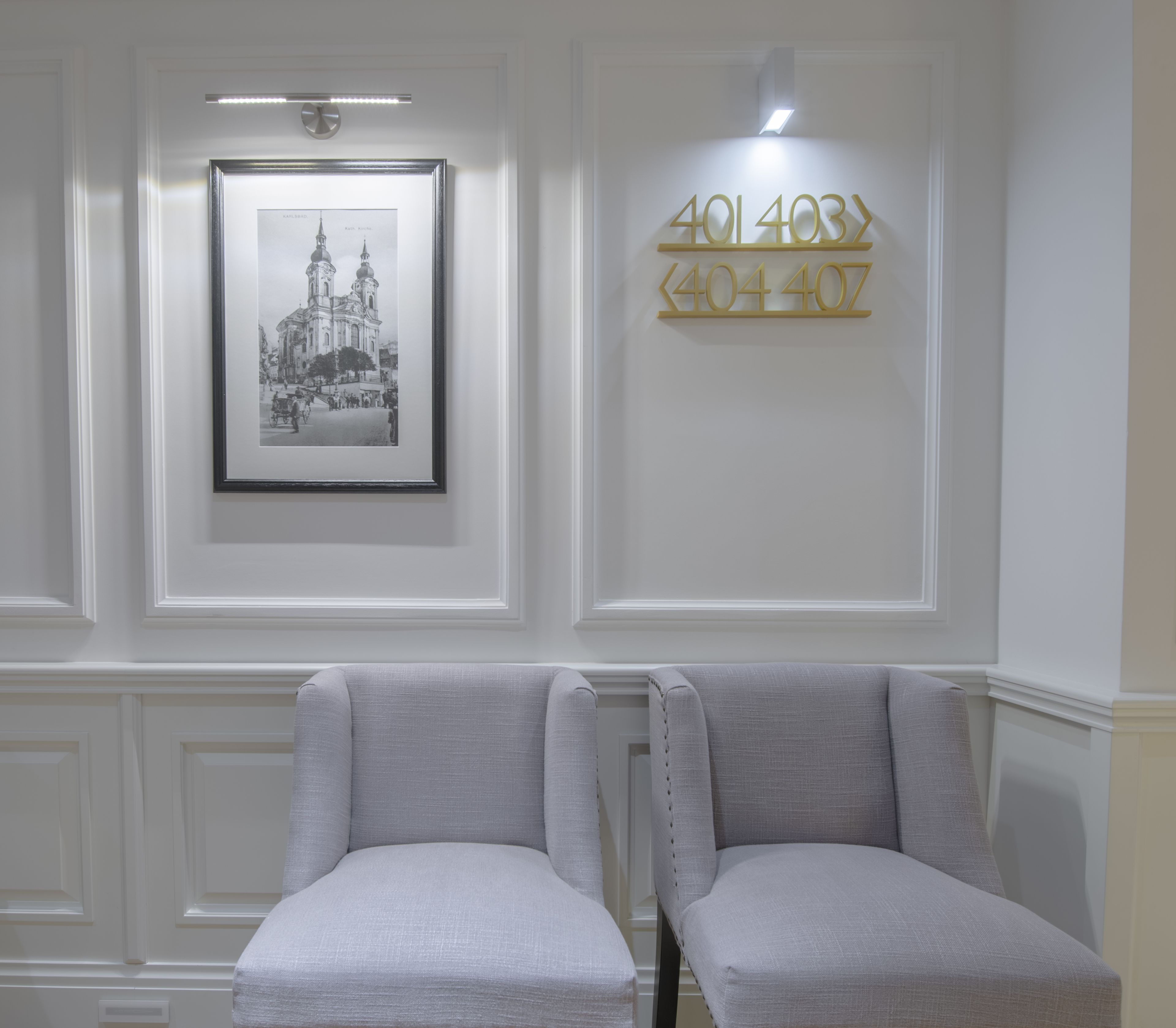 Una habitación con dos sillones grises tapizados, una fotografía en blanco y negro de un edificio histórico enmarcada, y un letrero con números colgado en la pared.